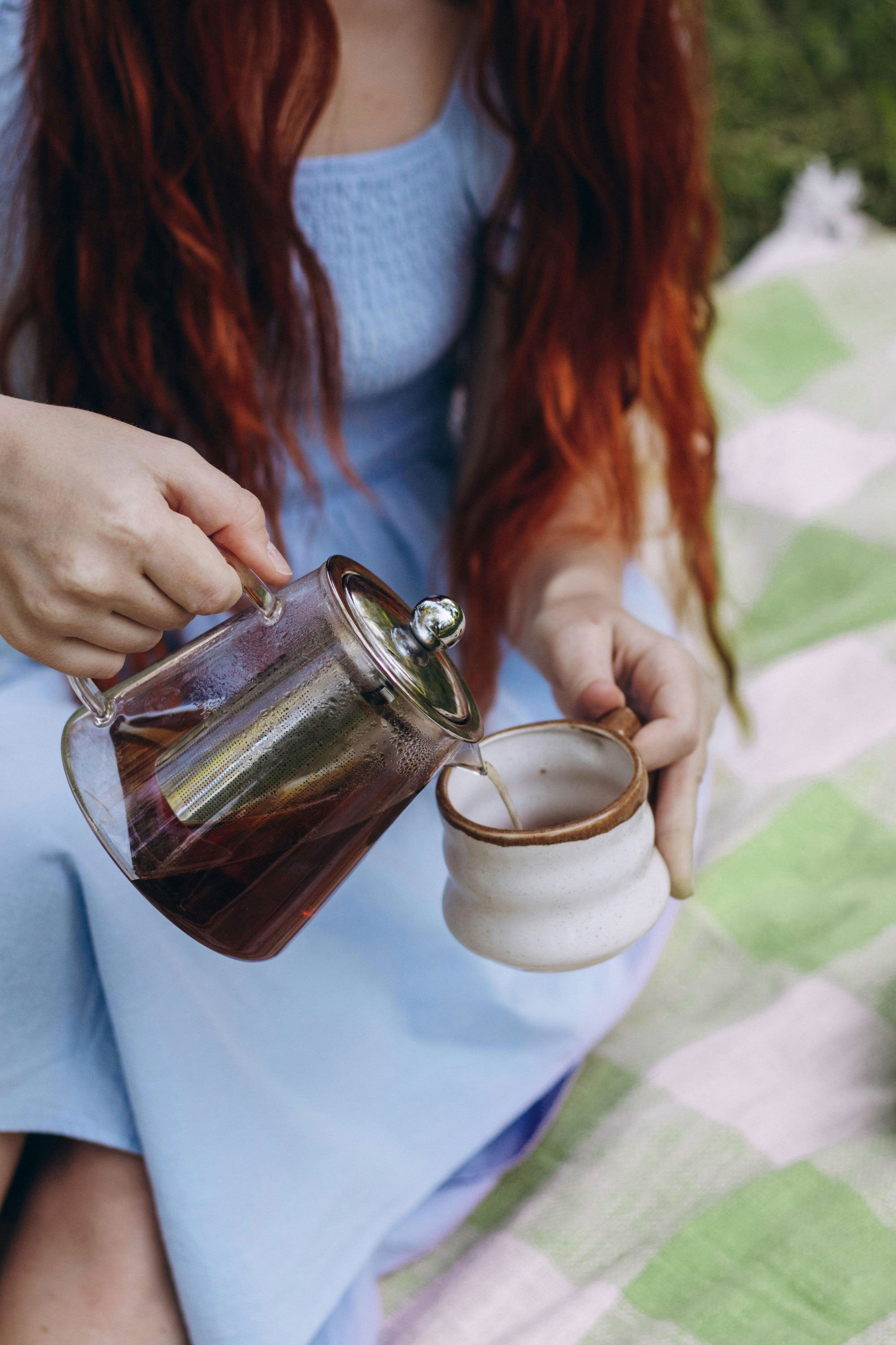 Loose leaf tea being poured from a glass infuser teapot into a ceramic mug outdoors, picnic setting